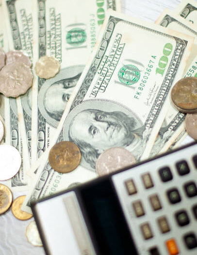 Money currency exchange coins penny dime over the hundred dollar bank notes on the white particle board table top diagonal view with vintage retro small calculator foreground defocus