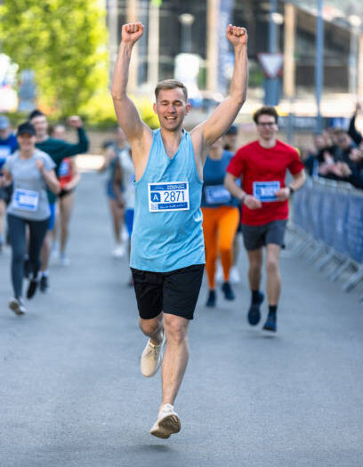Athletic Young Man Crossing the Finish Line in a City Marathon During the Day. Resilient Winning Runner Celebrating While his Family and Friends Cheer for him in the Audience